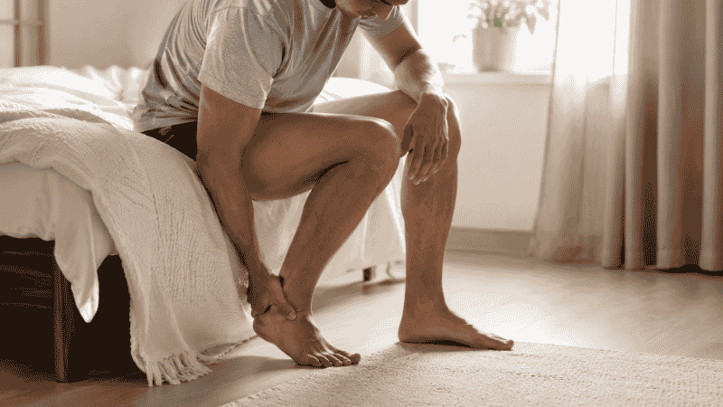 Person sitting on the edge of a bed in the morning holding their heel in pain before taking their first steps, representing the classic plantar fasciitis morning heel pain symptom