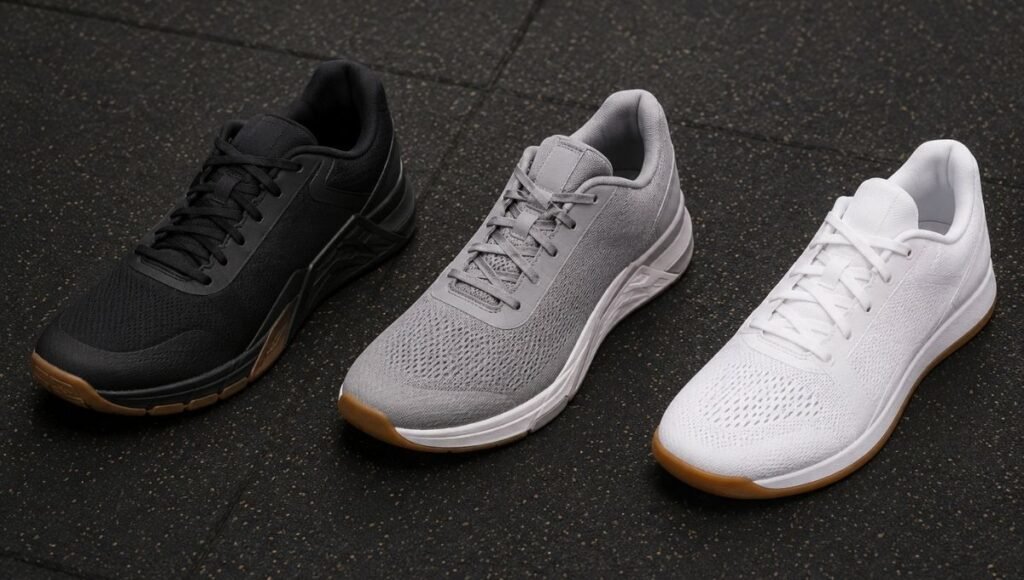 Three CrossFit training shoes placed side by side on a gym rubber floor, representing the Nike Metcon, Reebok Nano, and NOBULL Trainer comparison for women doing CrossFit and mixed gym training